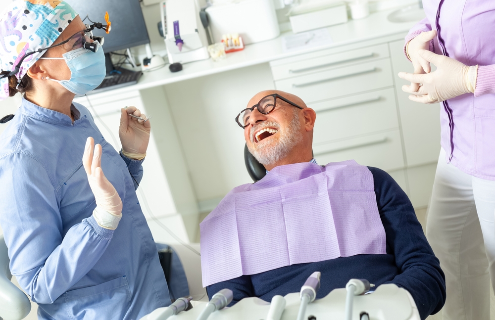 A happy family of four is at the dentist's office, with the mother holding a brochure titled "Understanding Buy-Up Dental Plans" and smiling reassuringly at her children. The dentist is pointing at an x-ray on the computer screen. No text on the image.